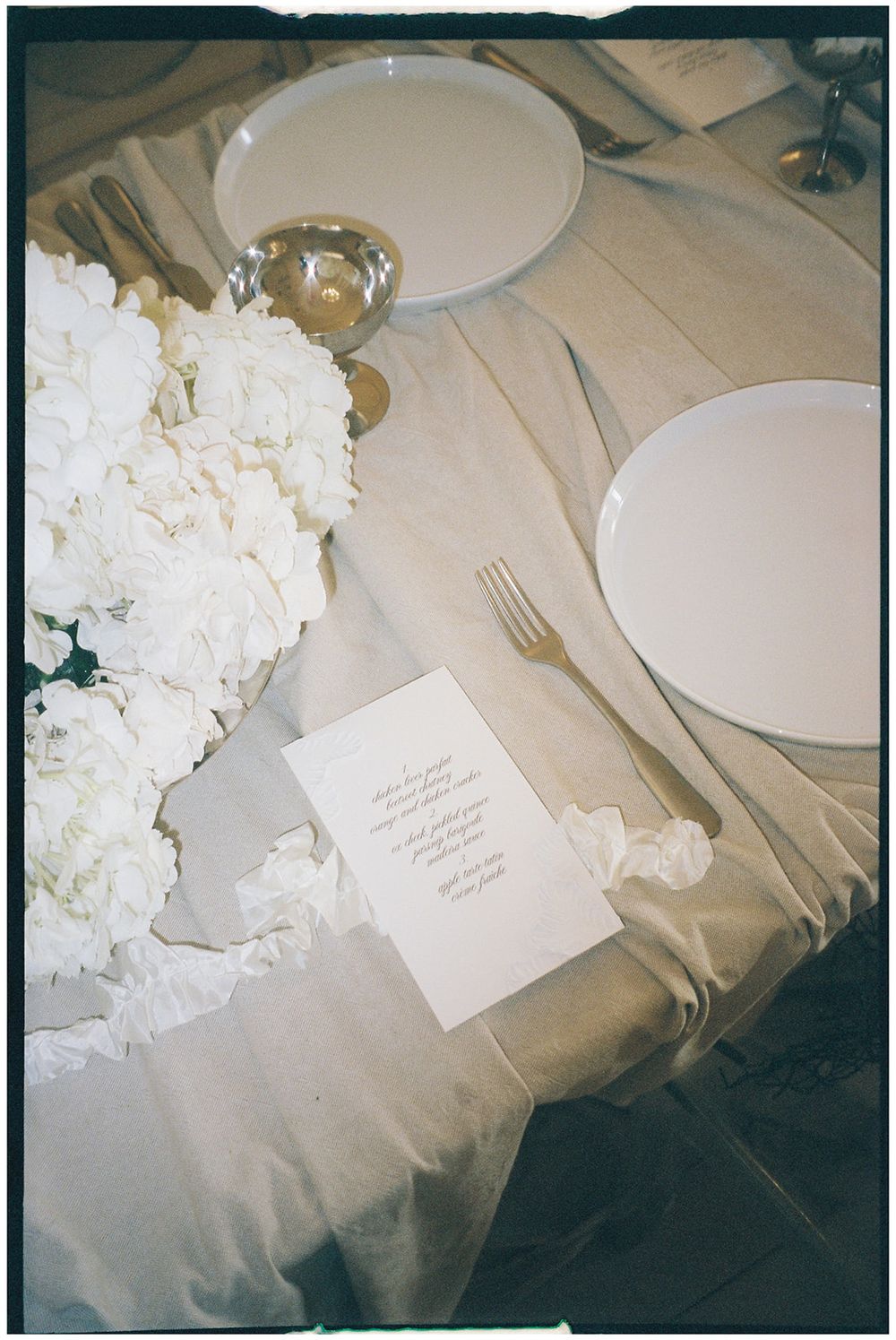 An elegant table setting with white hydrangeas, empty plates, and a small paper menu card on a beige tablecloth.