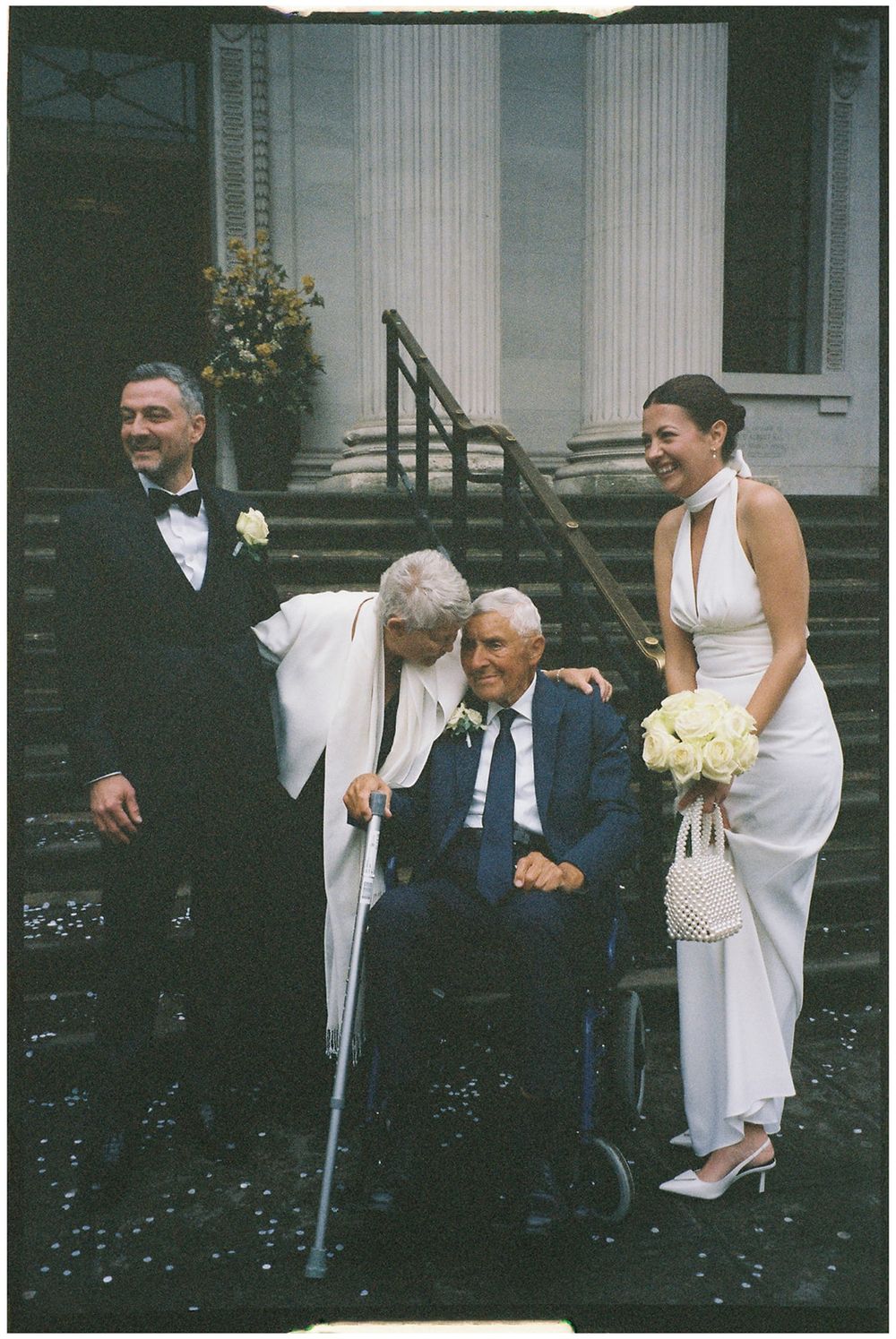 A group in formal attire gathers on stone steps with white columns in the background, with one person using crutches.