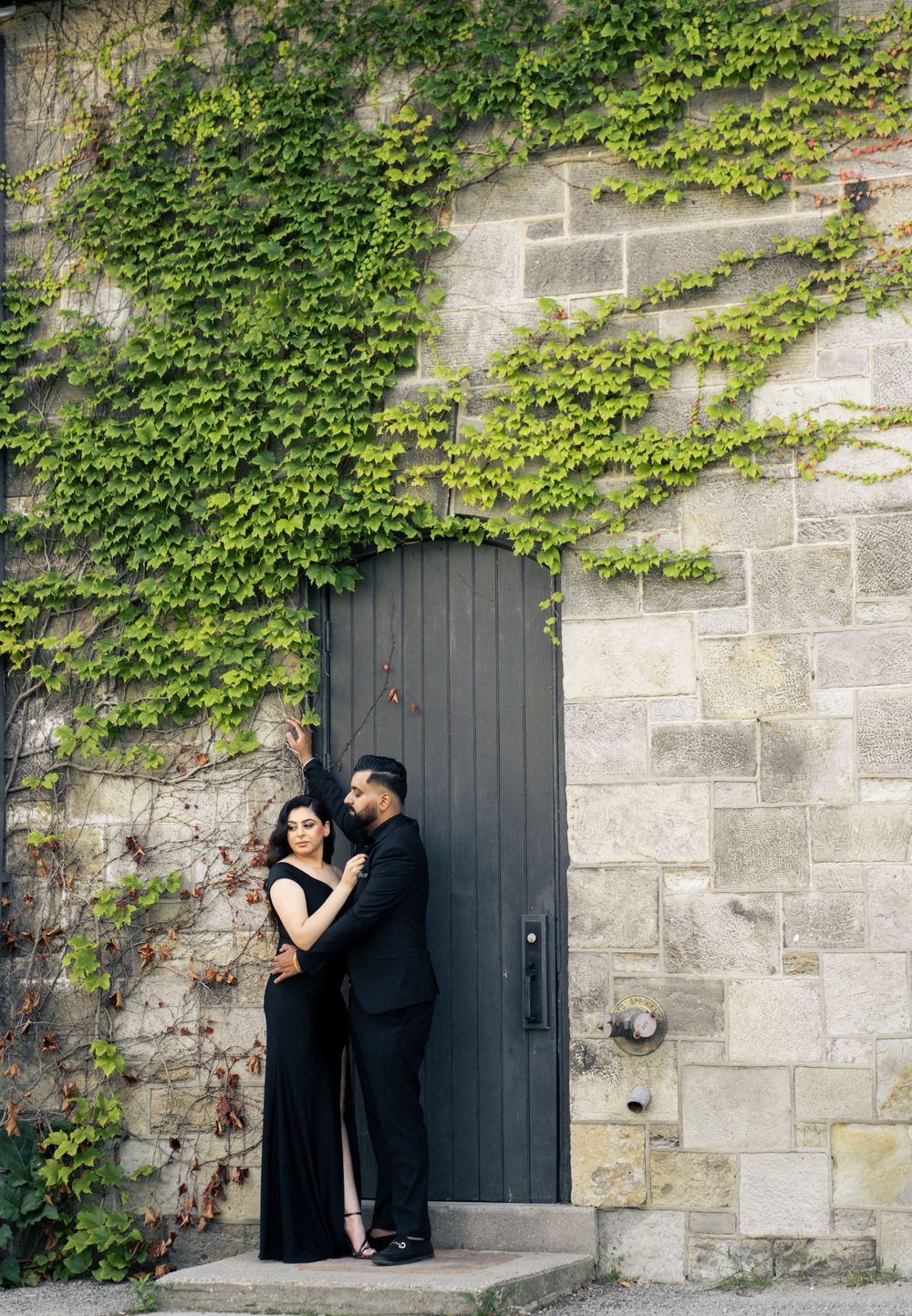 A romantic couple poses by an ivy-covered stone wall with black door.