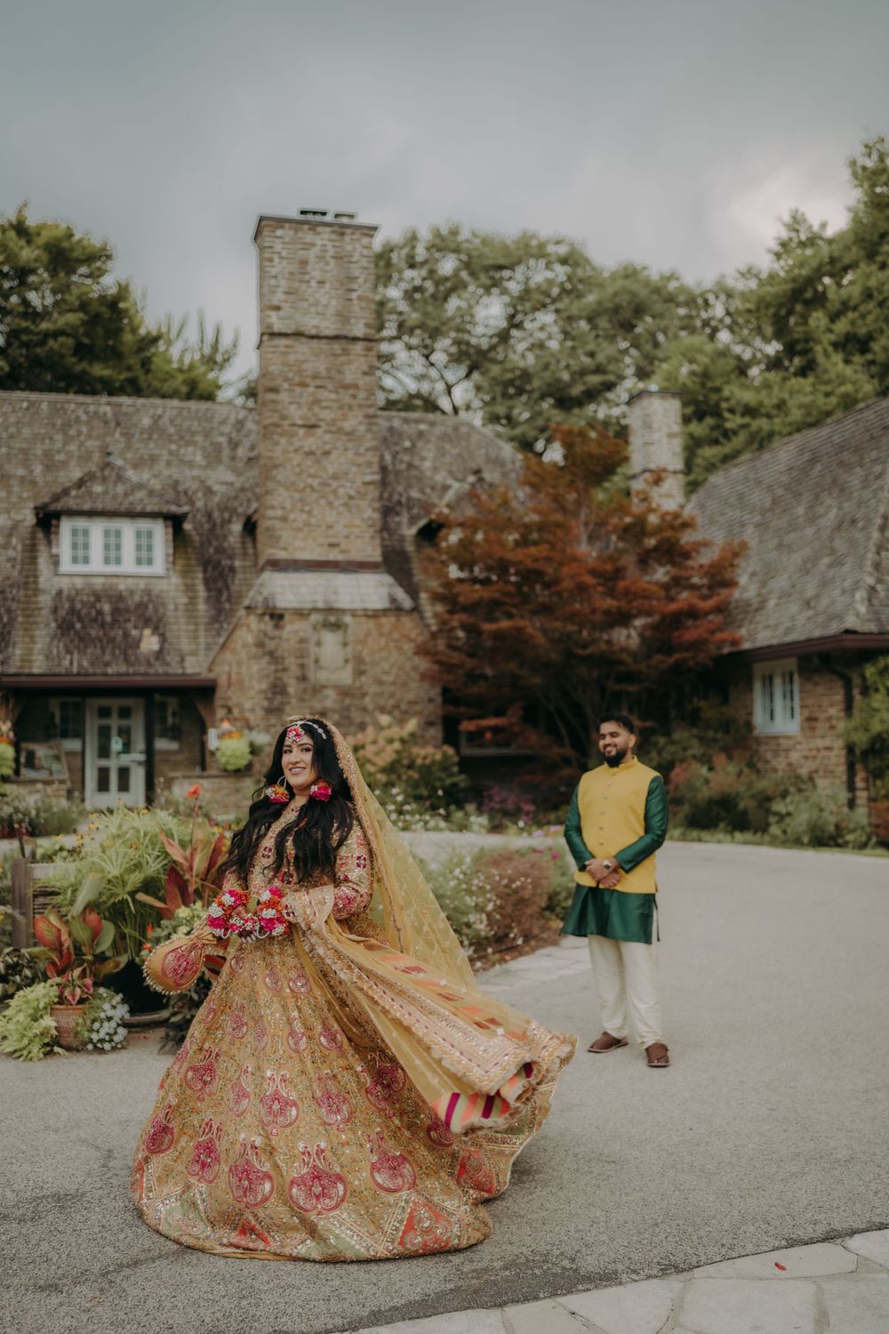 A couple in traditional Indian wedding attire poses in front of a stone cottage with autumn foliage.