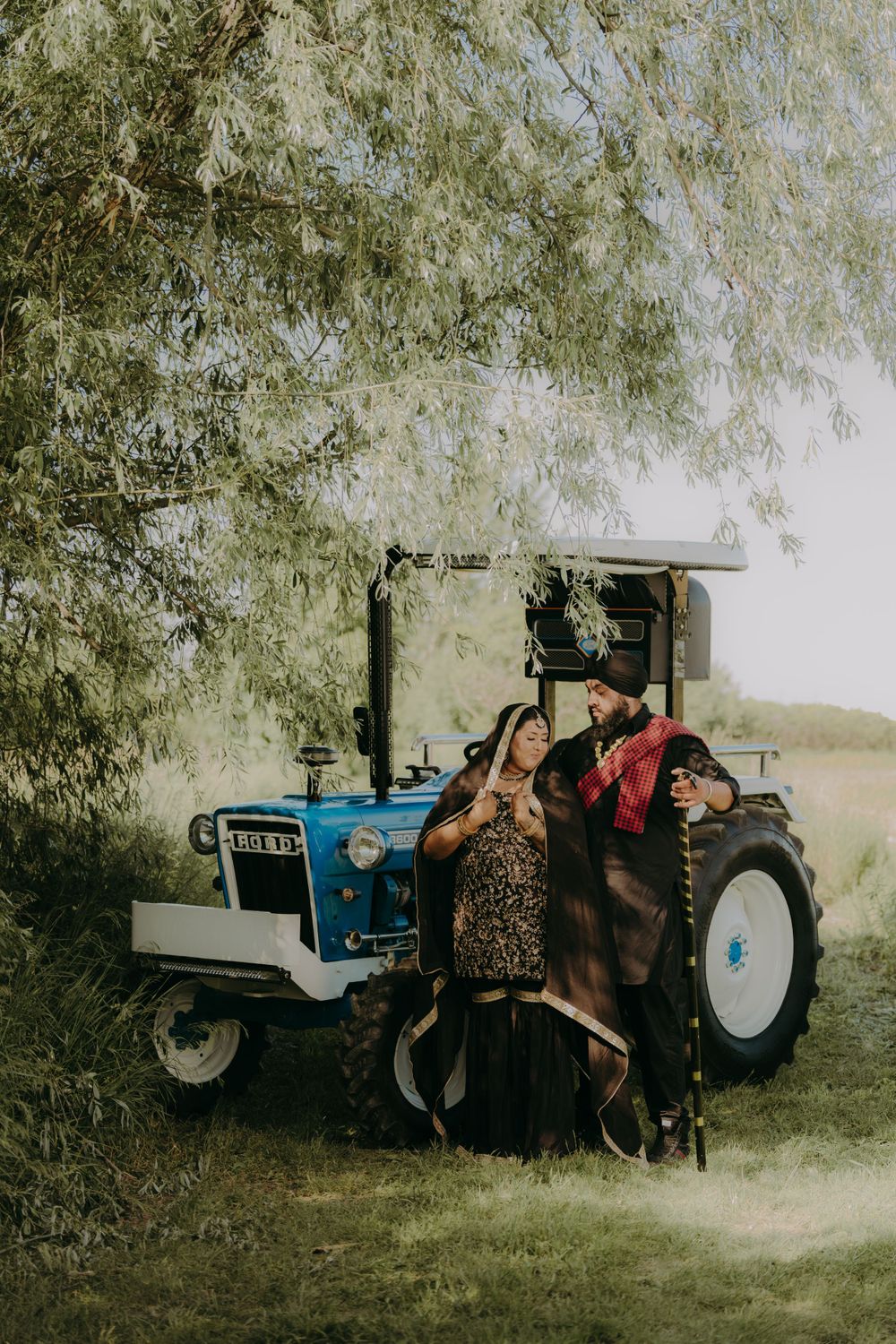 Couple posing playfully by vintage blue tractor under drooping tree branches.
