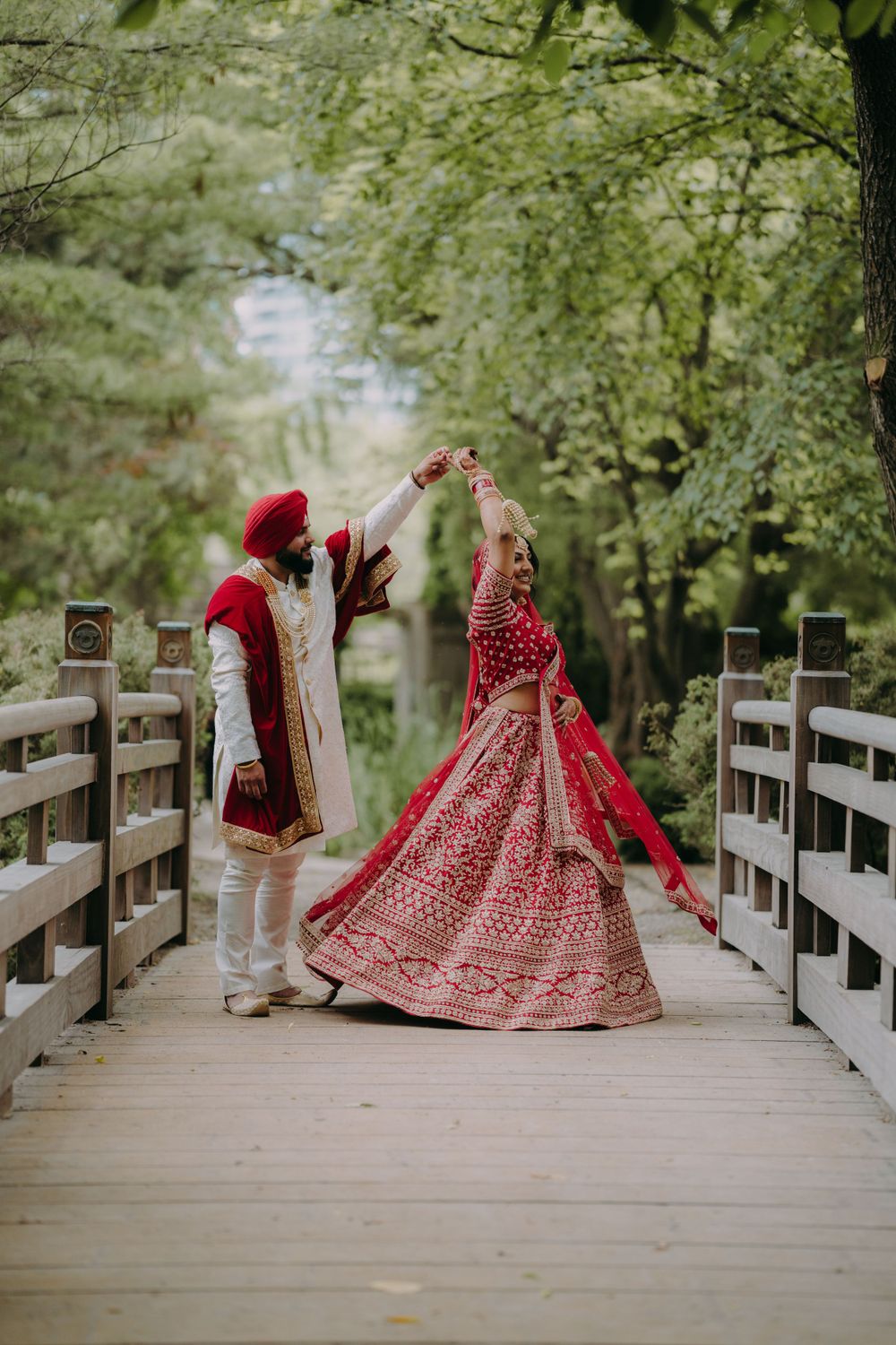Couple in traditional red Indian wedding attire pose joyfully on wooden bridge surrounded by greenery.