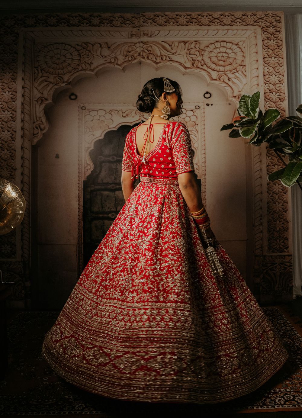 Ornate red and gold lehenga shown from behind against intricately carved archway in traditional setting.