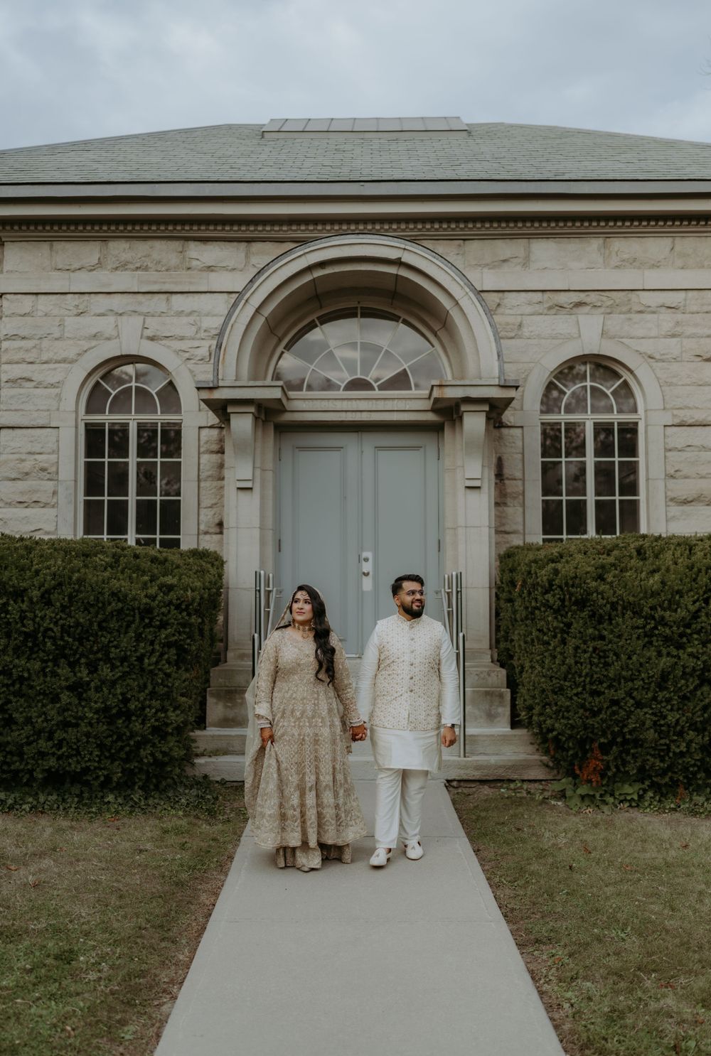 Couple in traditional Indian wedding attire stands in front of elegant stone building with arched doorway and windows.