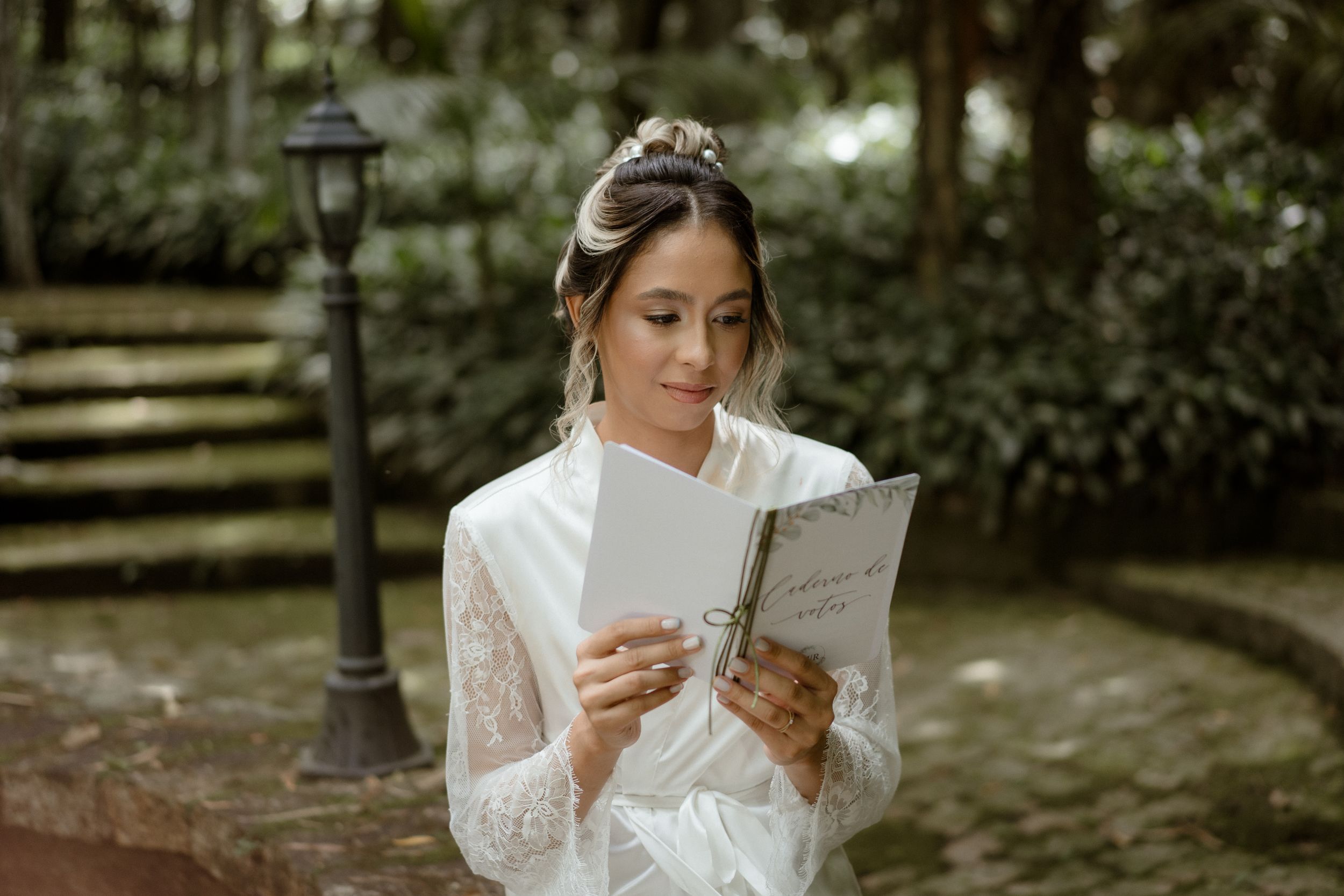 A person in white bridal attire holds a book while standing near a garden lamppost.