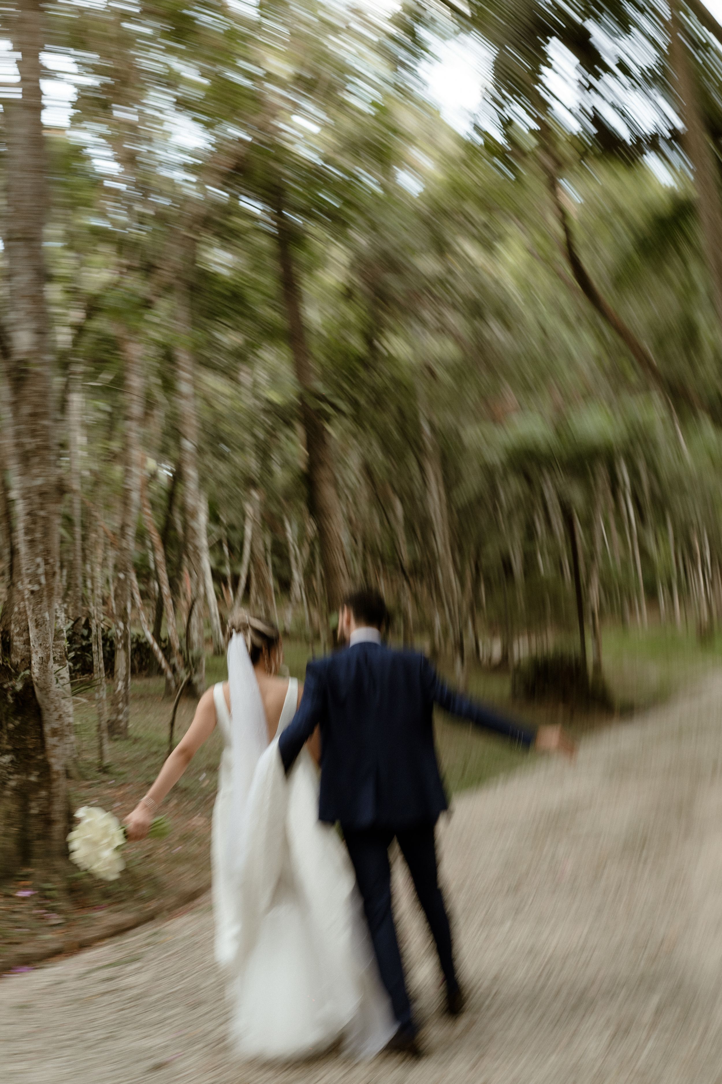 A wedding couple walking away down a forest path with pine trees blurred in motion effect.