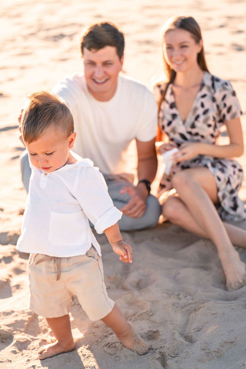 young boy walking on sand at beach while parents watch