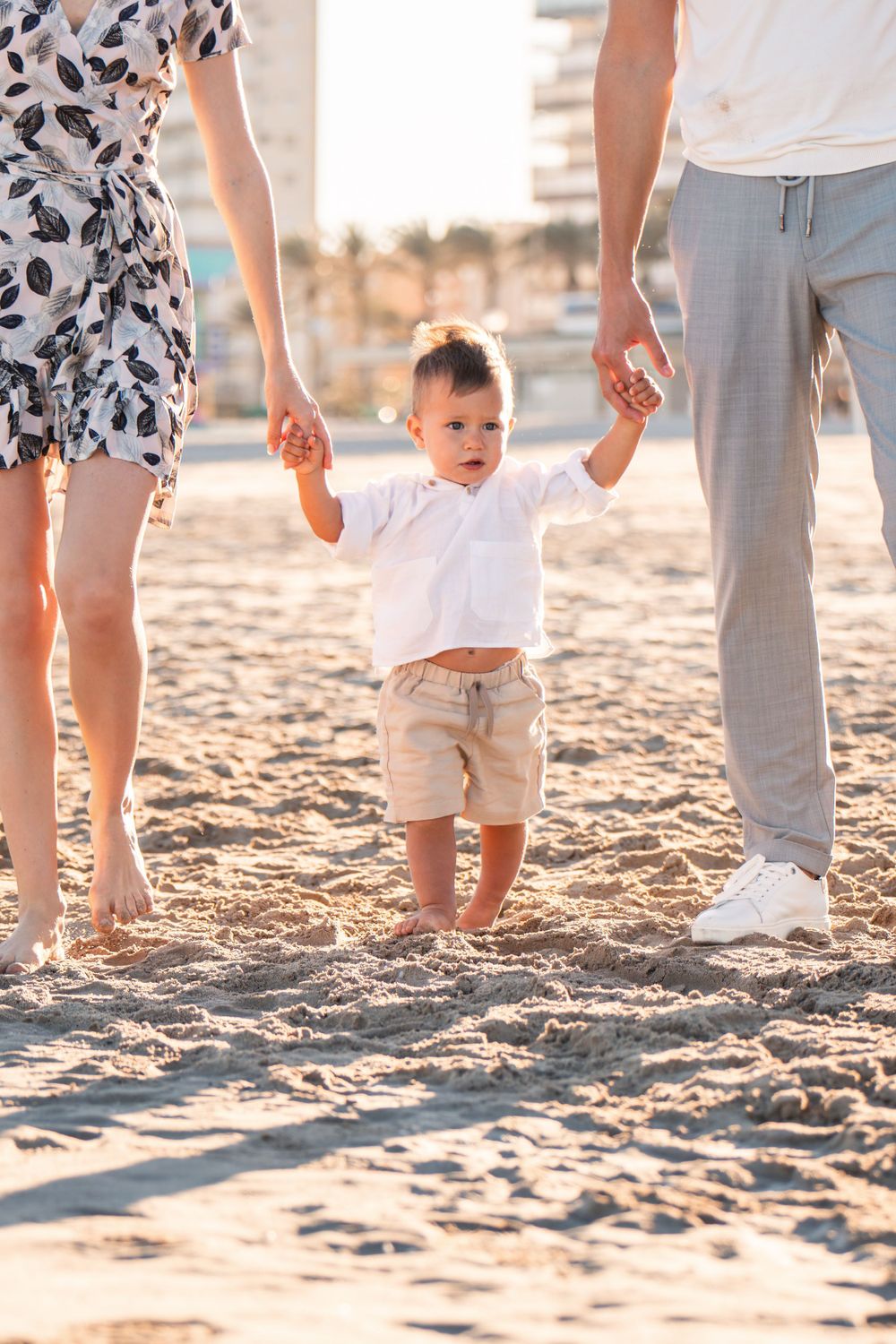 mom and dad both holding baby's hands to help him walk in the middle of them on the beach