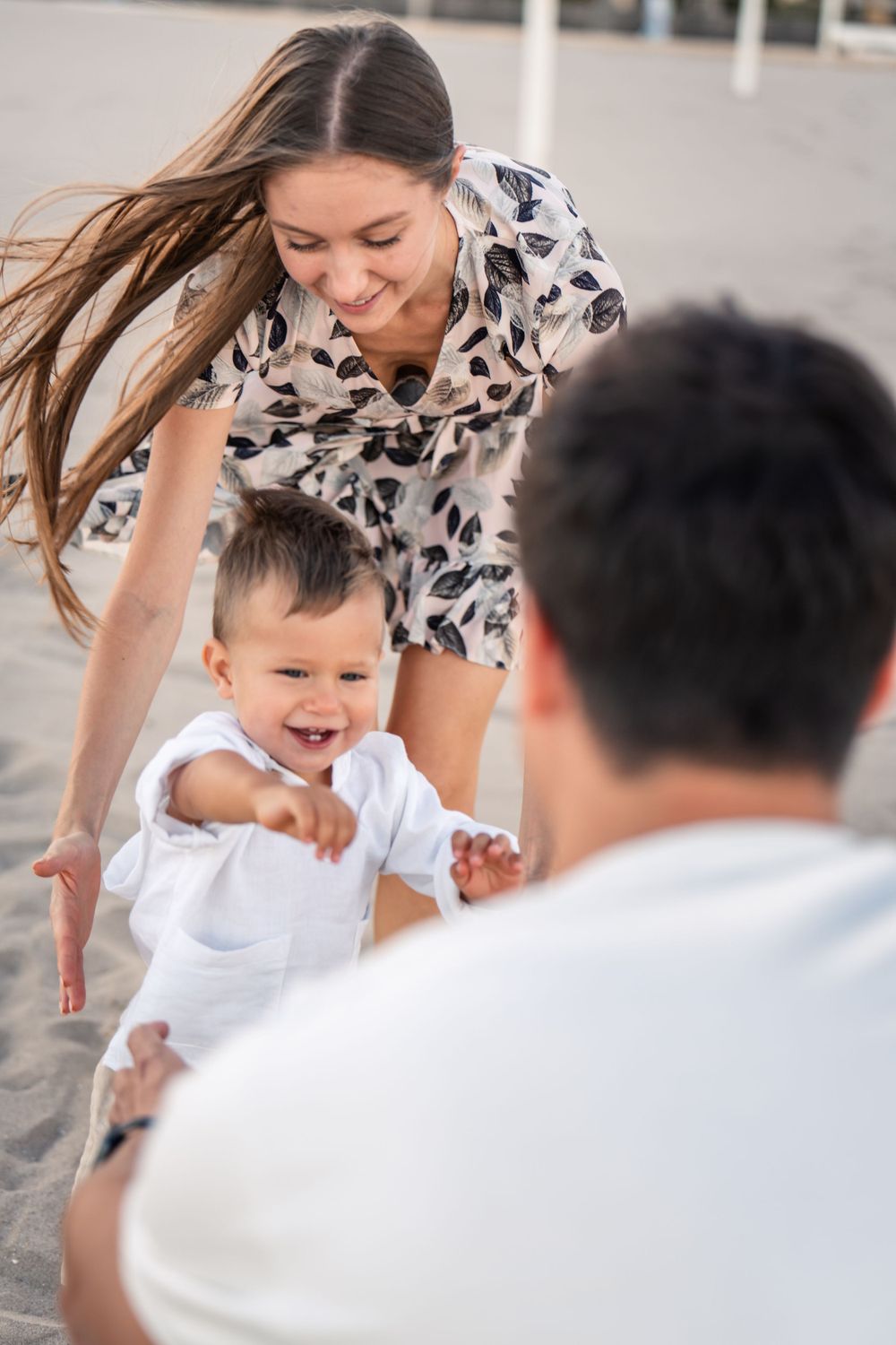 young boy walking towards dad with a big smile on his face while mom helps him walk