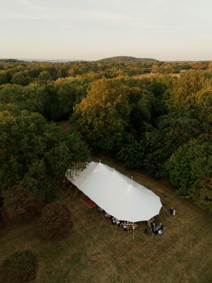 An aerial view shows a white event tent nestled among autumn trees in a clearing during golden hour.