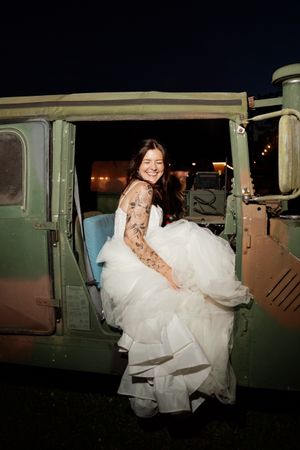 Smiling bride in white tulle gown with sleeve tattoos stepping out of a vintage military vehicle at night.