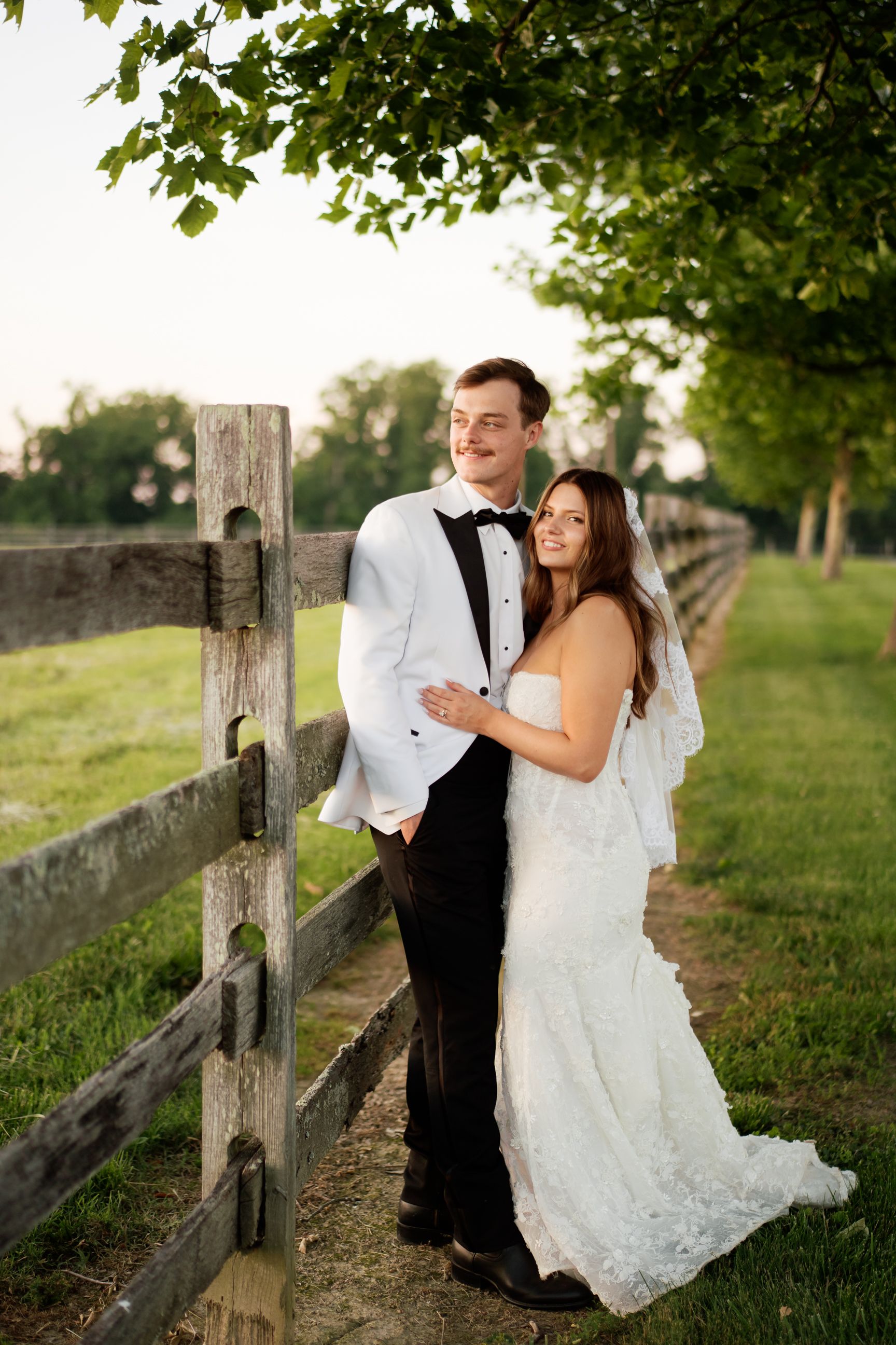 Bride and groom posing by a rustic wooden fence in a lush green outdoor setting during golden hour.