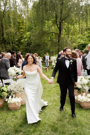 Newlywed couple walking down outdoor wedding aisle surrounded by white flowers and guests on a lush green lawn.