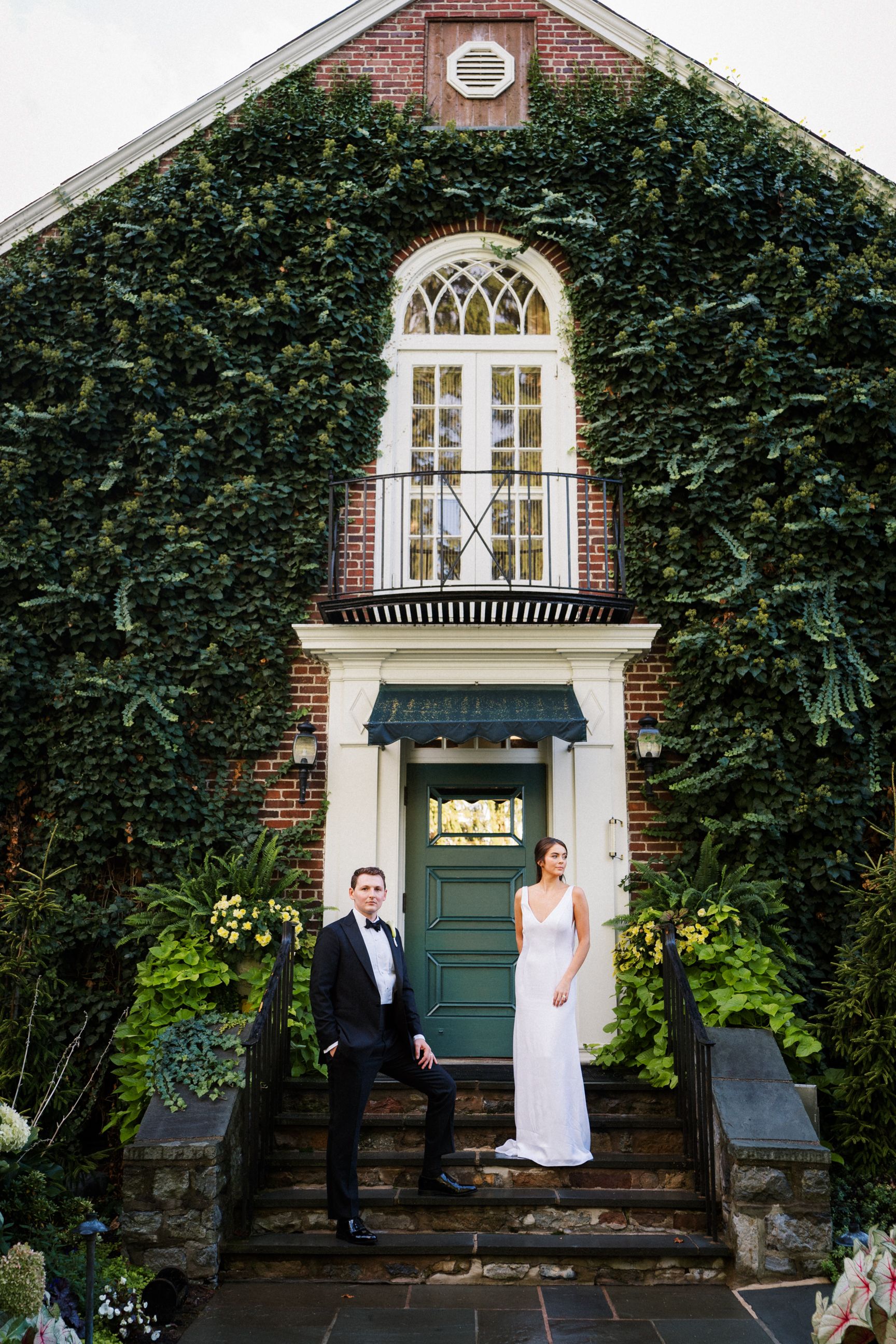 Elegant couple standing on stone steps in front of ivy-covered brick building with arched windows and green door.
