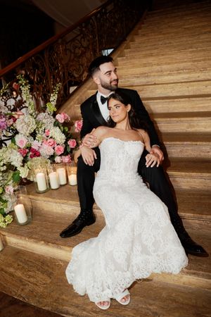Bride in lace gown and groom in tuxedo sit together on marble staircase with candles and pink floral arrangements.