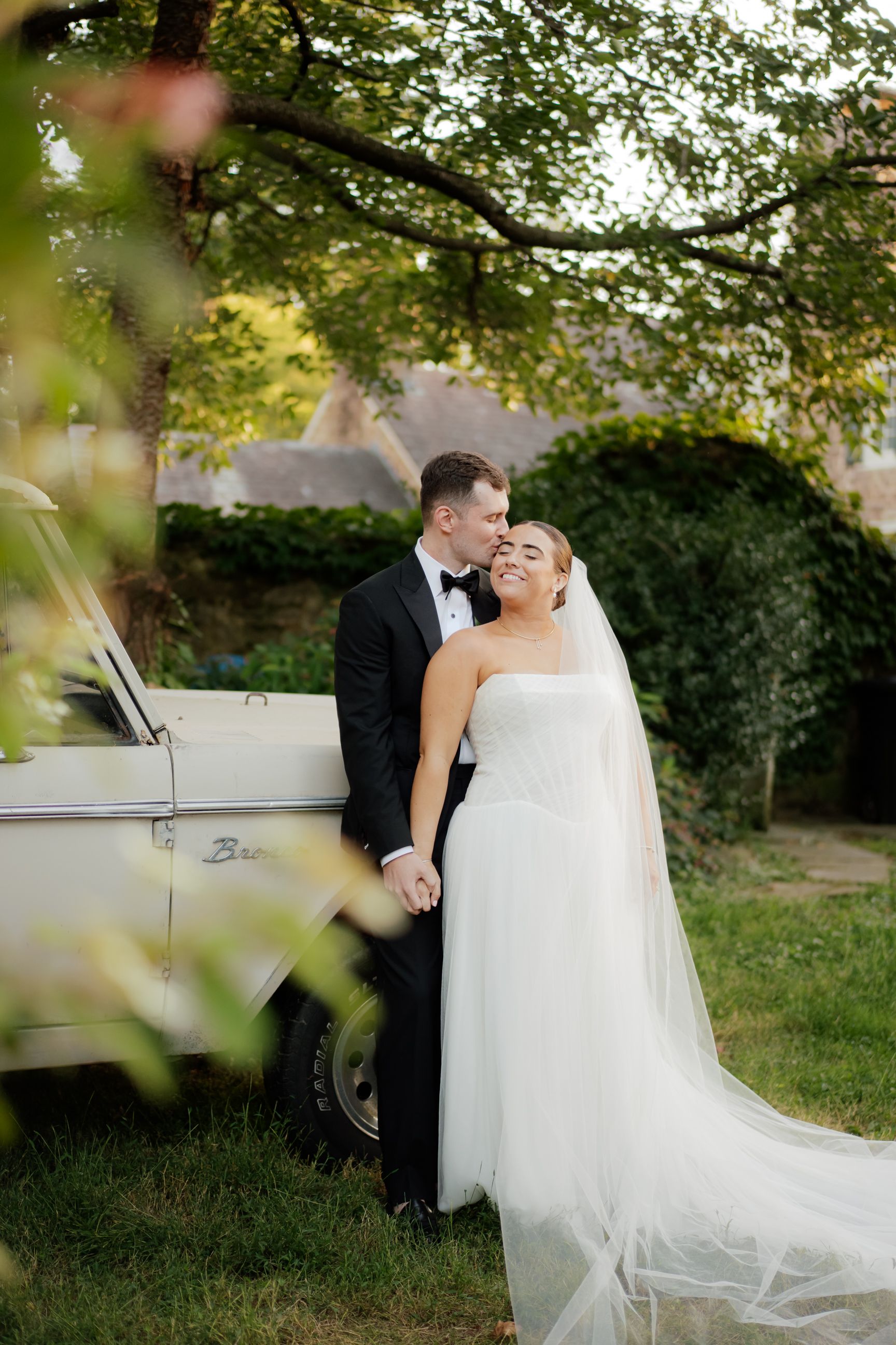 A romantic wedding couple embraces near their vintage car in a garden setting with natural greenery and soft lighting.