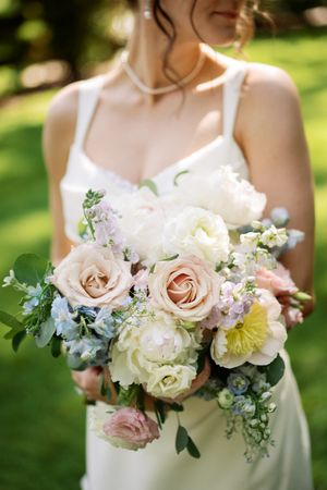 A romantic bridal bouquet featuring blush pink roses, white peonies, and delicate greenery held outdoors in natural lighting.