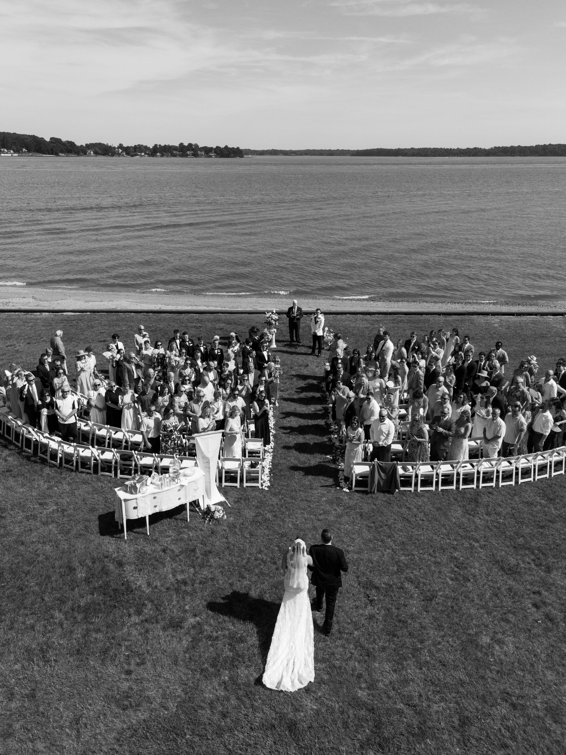 Aerial black and white photo of a wedding ceremony on a waterfront lawn with guests seated in a semicircle.