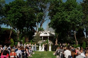 Outdoor garden wedding ceremony with white gazebo surrounded by tall trees and guests seated on the lawn.