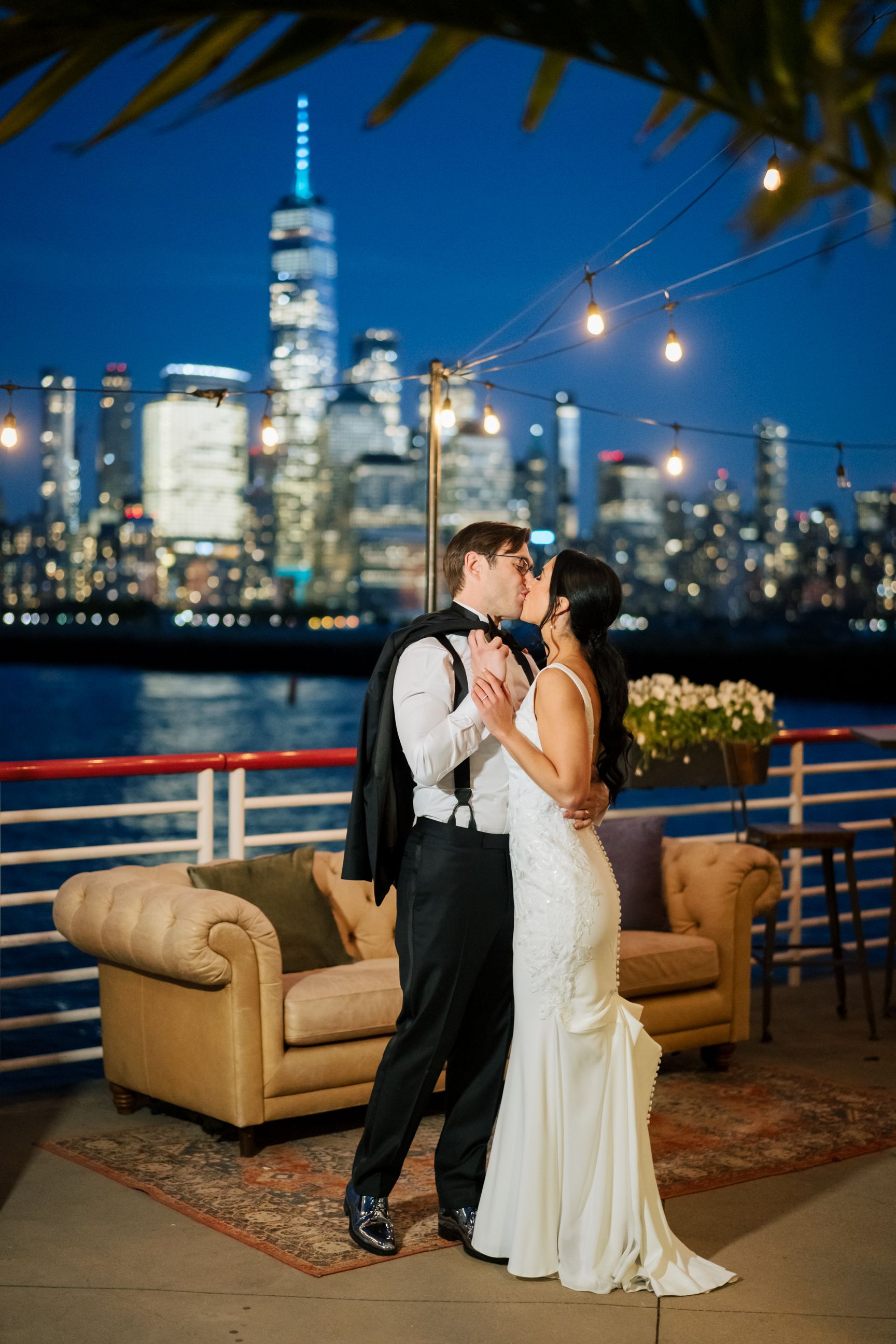 A romantic nighttime wedding portrait with the New York City skyline and string lights in the background.