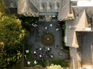 Aerial drone view of people gathered in line at historic European castle entrance under late afternoon sun.