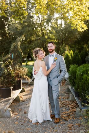Elegant couple in wedding attire pose on garden path with golden sunlit trees in background.