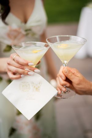 Two hands holding elegant martini glasses with lemon garnished cocktails and a white cocktail napkin at a celebration.