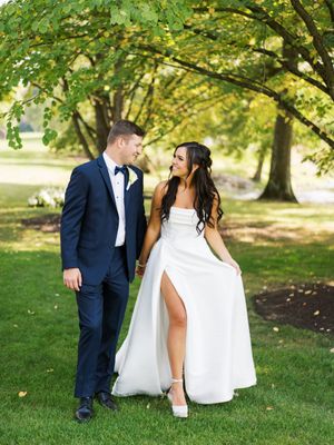 A romantic wedding photo of a couple smiling at each other under lush green trees in a garden setting.