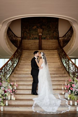 Bride and groom share a kiss at the base of an elegant double staircase adorned with candles and pink florals.