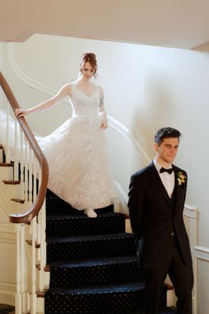 Bride in lace gown descends elegant staircase while groom in black tuxedo waits below during wedding first look.