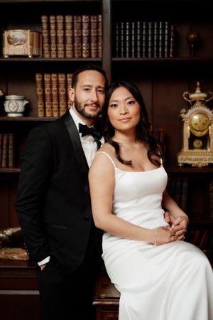 Elegant couple poses in formal attire against library backdrop with vintage books and antique clock decor.