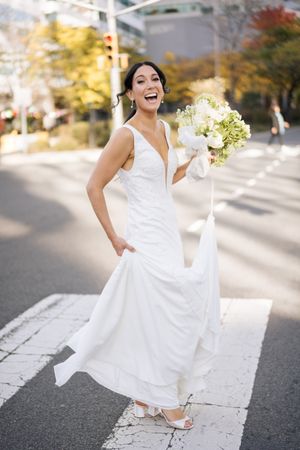 A radiant bride in a fitted white gown laughs while holding a bouquet on a city street.