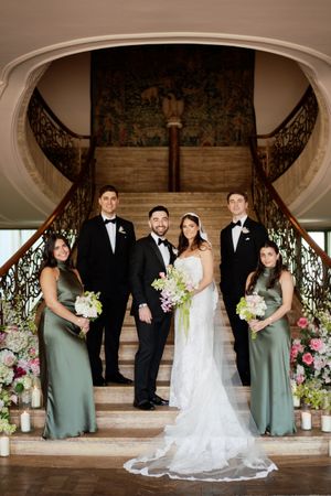 Wedding party poses on grand staircase, bride in lace gown with bouquet, groom in tuxedo, bridesmaids in sage green dresses.