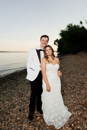 A couple in wedding attire poses on a rocky beach at sunset, groom in white tuxedo jacket, bride in strapless lace gown.