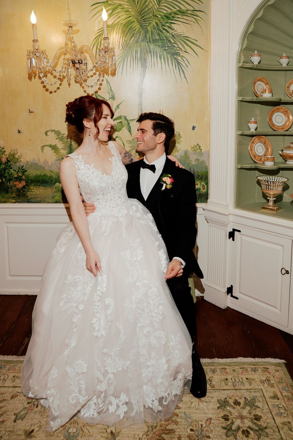 A bride in a lace ballgown and groom in a black tuxedo share a romantic moment in an elegant, chandelier-lit room.