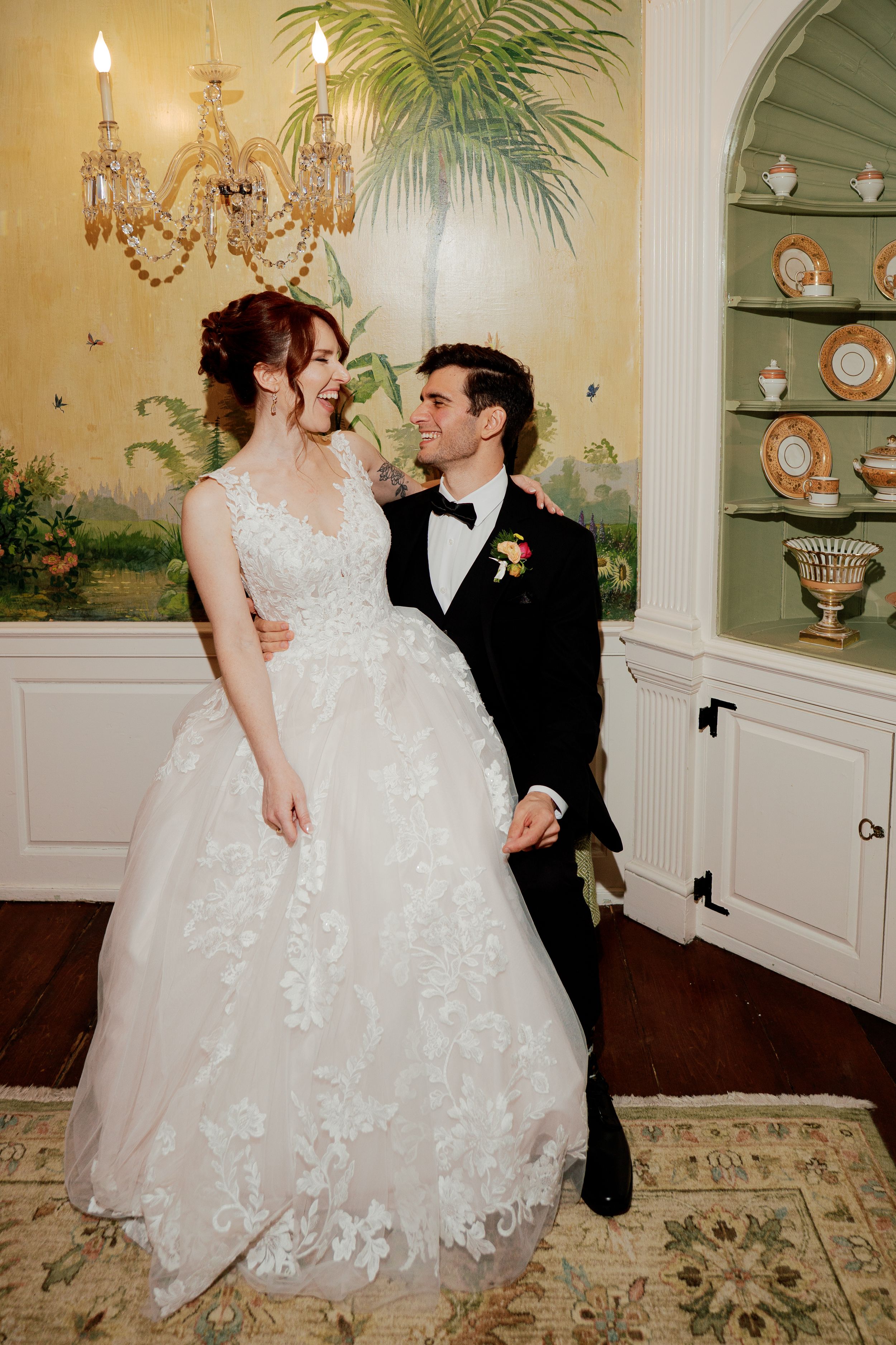 A bride in a lace ballgown and groom in a black tuxedo share a romantic moment in an elegant, chandelier-lit room.