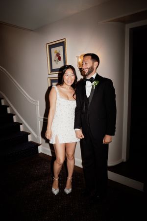 A couple poses together at a wedding reception, with her wearing a short white dress and him in a black tuxedo.