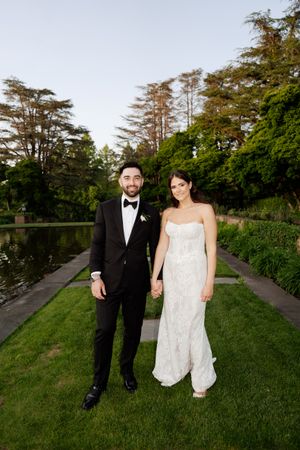 A bride and groom hold hands smiling in a lush garden, she in a white gown, he in a black tuxedo.