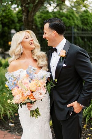 Joyful bride and groom share a candid moment while holding coral and peach floral bouquet in garden setting.