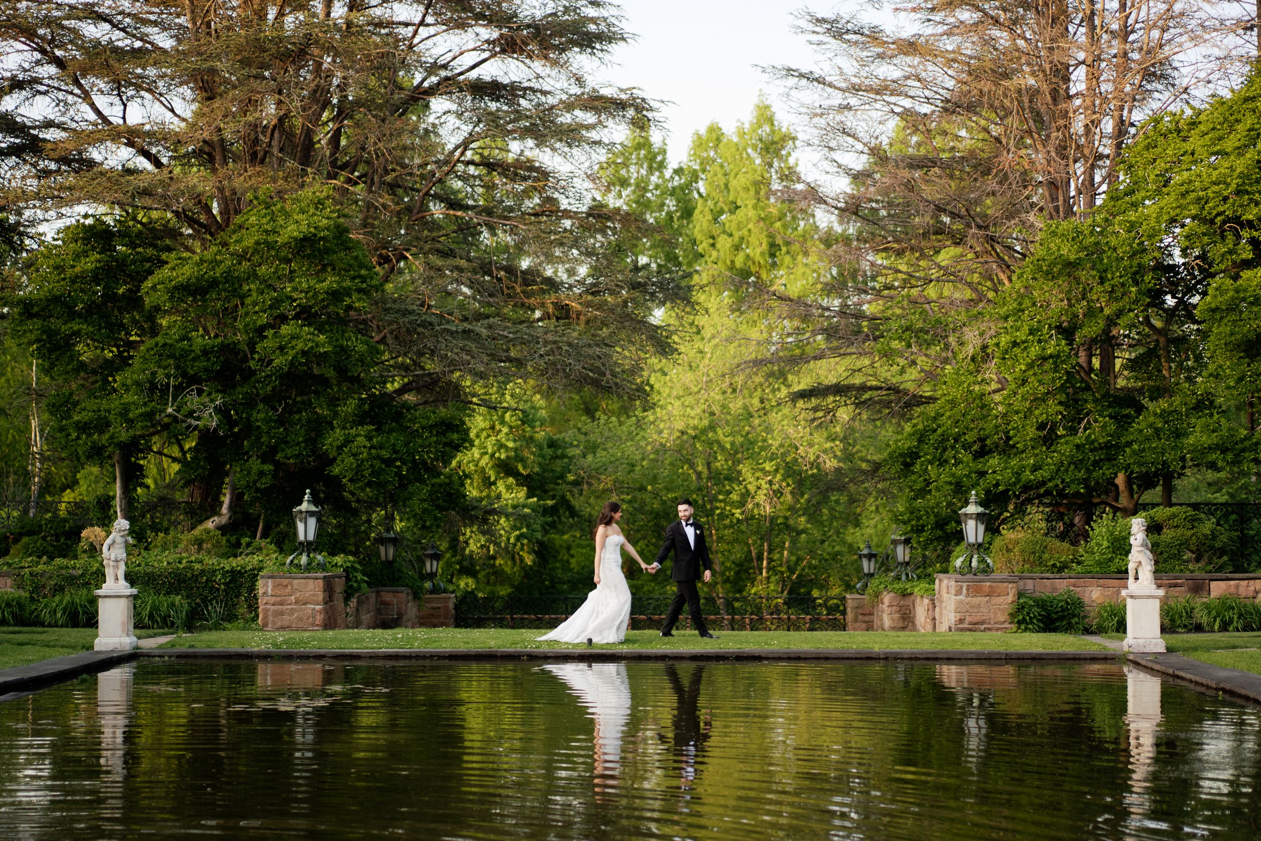 Bride and groom walk hand in hand beside a reflective garden pond surrounded by lush green trees.