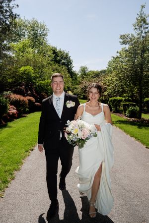 Newlyweds walk together on stone path through lush garden landscape on sunny wedding day.