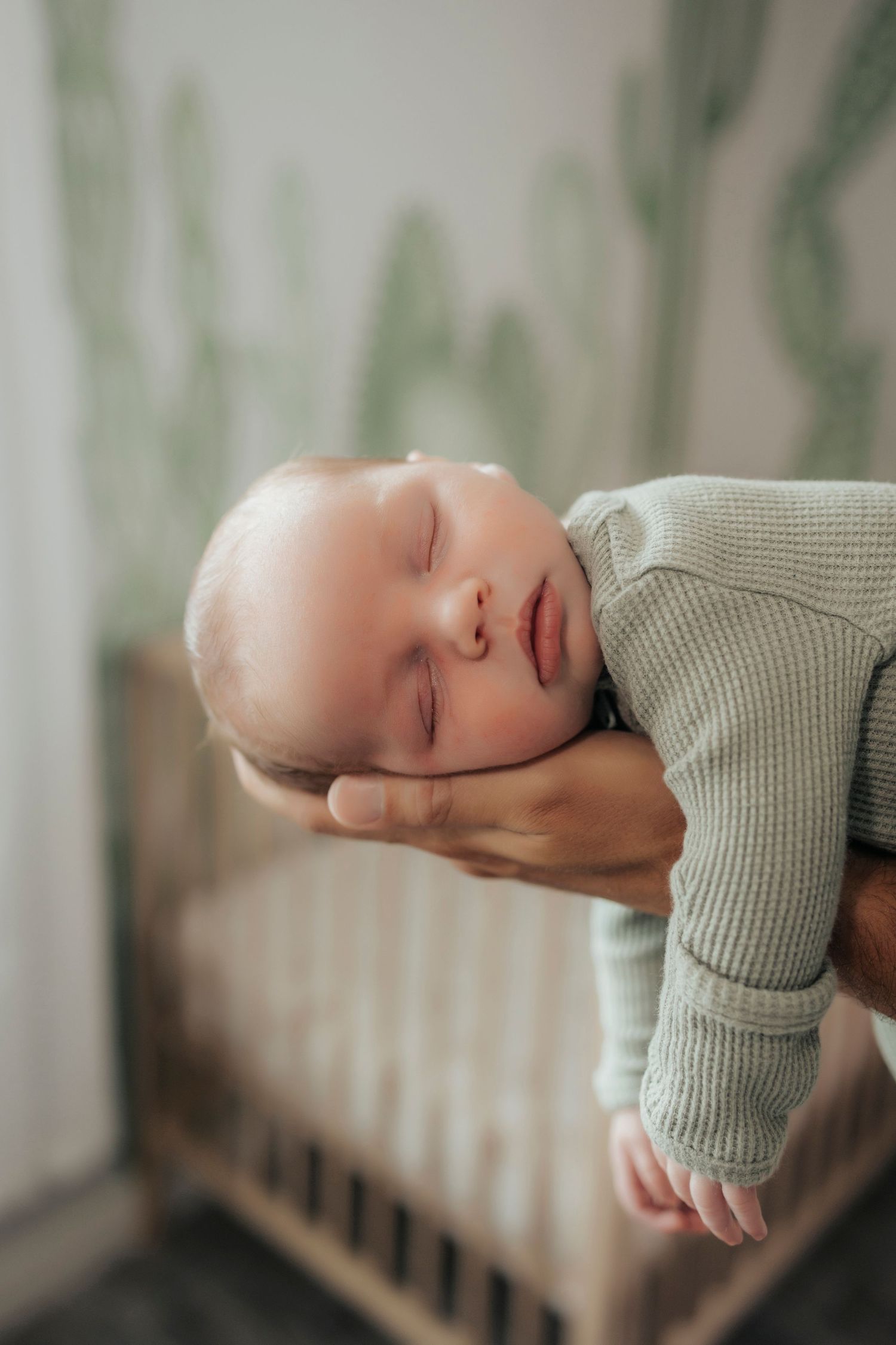 A sleeping infant rests peacefully on a supportive hand near a crib.