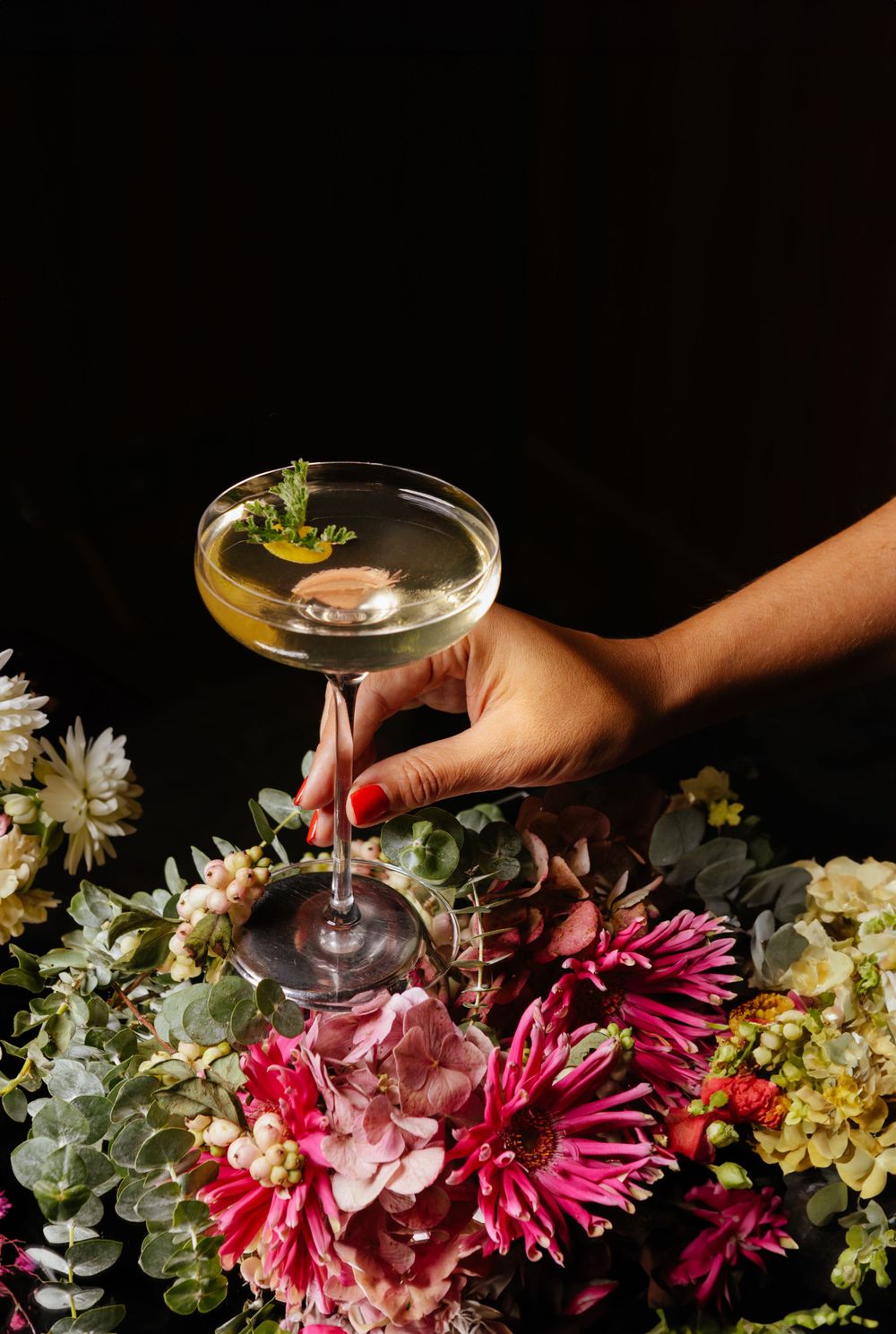 Hand holding martini glass over colorful floral arrangement with pink and red blooms.