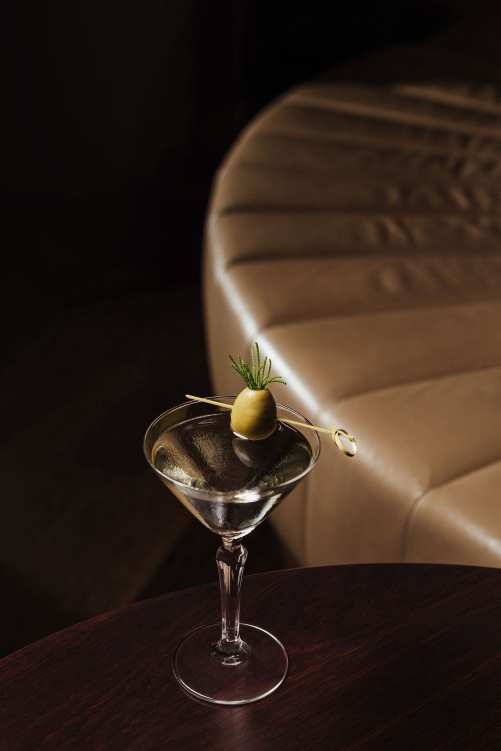 Martini glass with olive garnish photographed against leather booth seating.