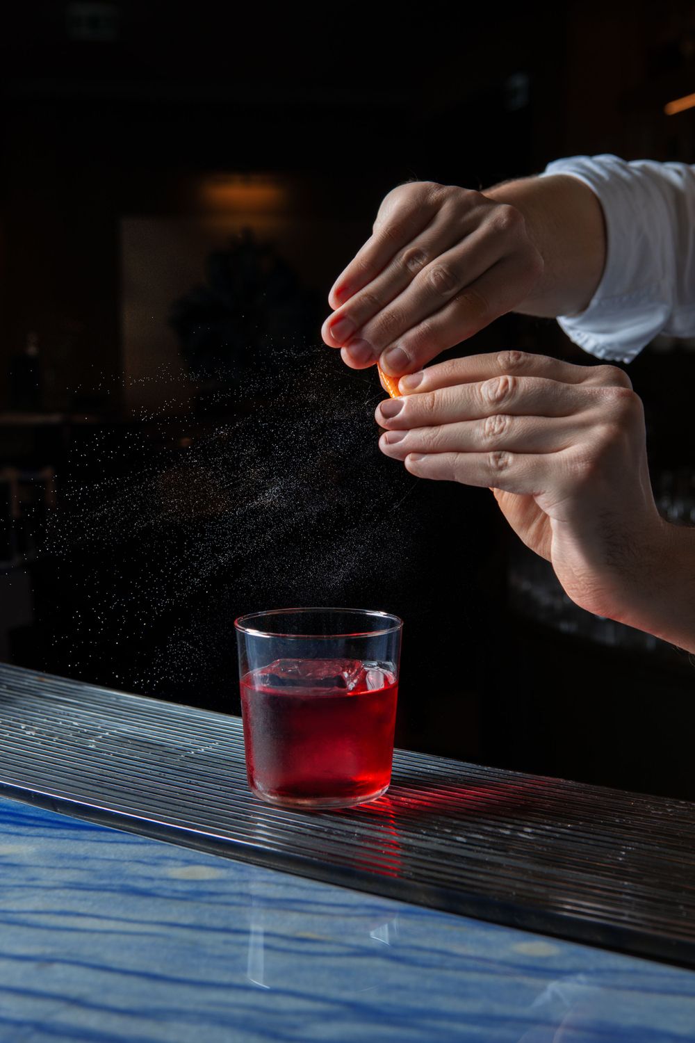 Hands sprinkling garnish over red cocktail in dramatic lighting.