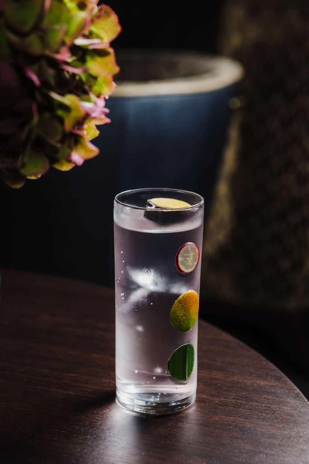 Tall glass of clear cocktail garnished with citrus slices beside purple hydrangea flowers.