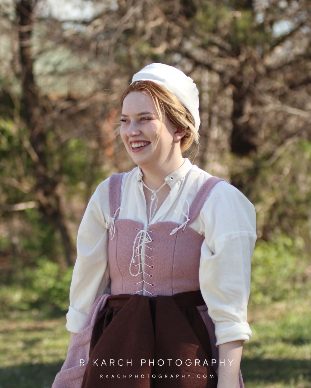 A historical period photoshoot in a garden setting features someone in a pink corset dress and white bonnet.
