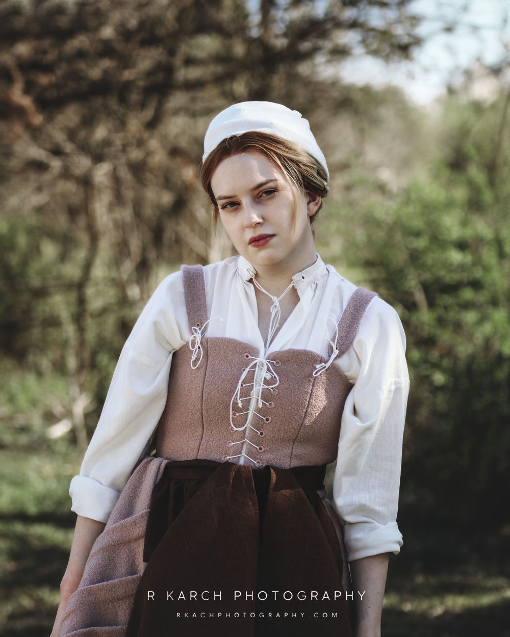 Historical costume portrait featuring brown corset dress and white cap in natural setting.