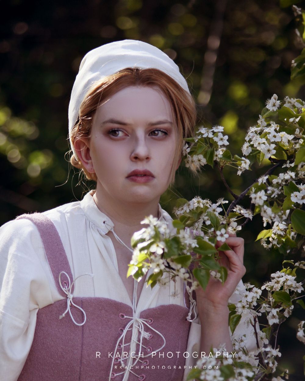 A portrait in historical dress shows someone amid spring blossoms wearing a pink corset and white cap.