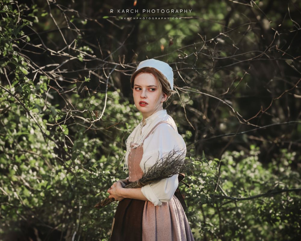 Period drama style portrait in rustic dress and cap against green foliage background.